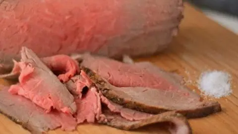 A roast beef roast with thin slices cut off in the front of it on a cutting board.