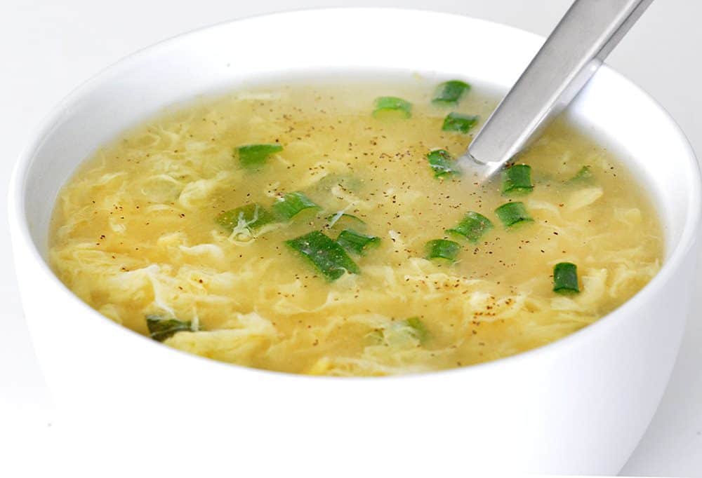 Close up of a silver spoon and white bowl containing egg drop soup.