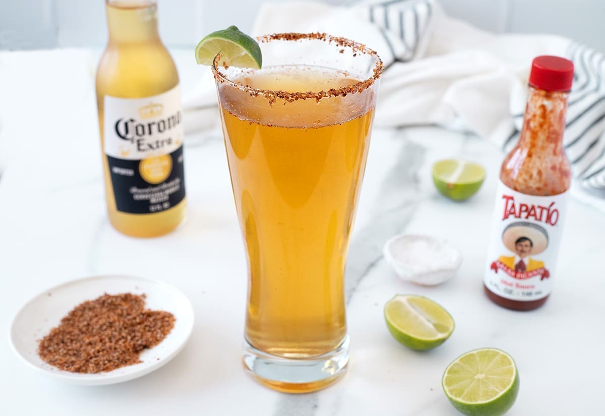Glass of michelada next to hot sauce and a bottle or corona.