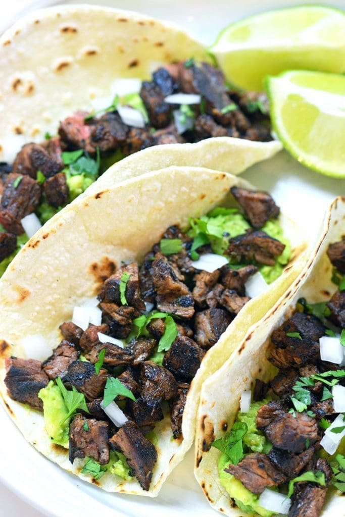 Three Carne Asada tacos topped with cilantro and onions on a white plate. Served with lime slices and cilantro.