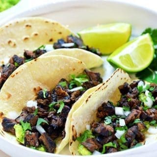 Three Carne Asada tacos topped with cilantro and onions on a white plate. Served with lime slices and cilantro.