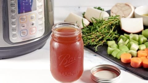 Picture of bone broth in a glass mason jar flanked by an Instant Pot machine and cut vegetables including carrots, celery, onions and garlic.