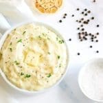 White bowl of garlic mashed potatoes on a white marble background. Roasted garlic, salt and pepper are next to the bowl.
