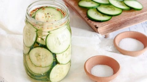 A glass mason jar full of sliced cucumbers and smashed garlic cloves to make picked cucumbers. Wooden cutting board with sliced cucumbers and bowl of sugar and salt in the background.