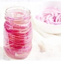 Pickled red Onions in a glass mason jar flanked by a bowl of cut red onions in a white bowl
