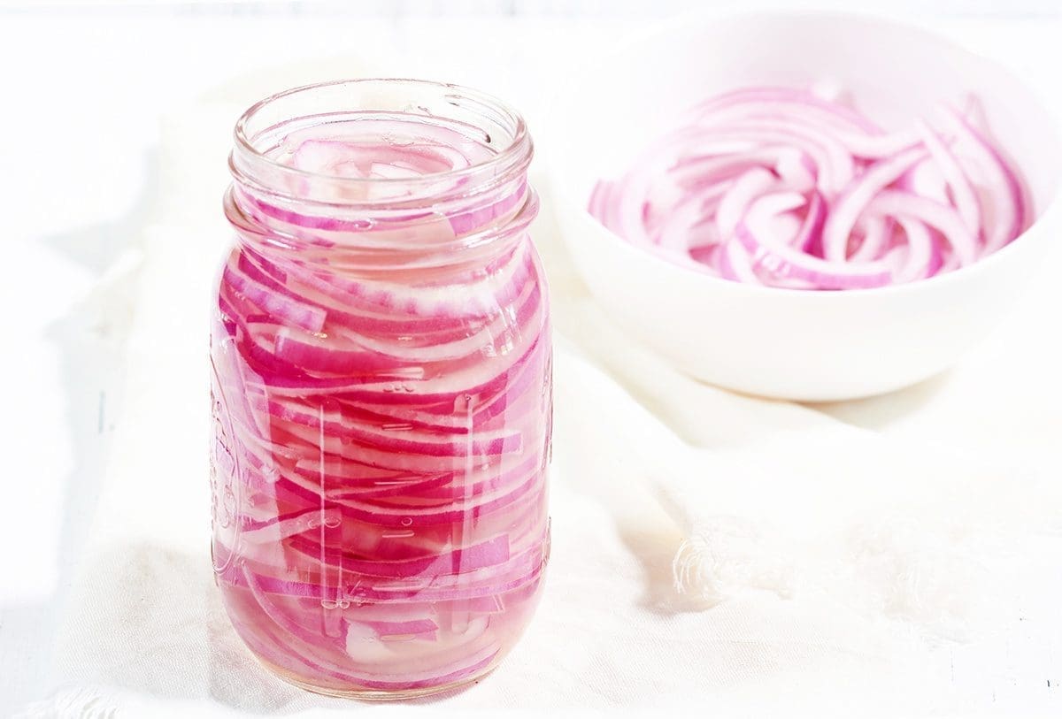 Pickled red Onions in a glass mason jar flanked by a bowl of cut red onions in a white bowl