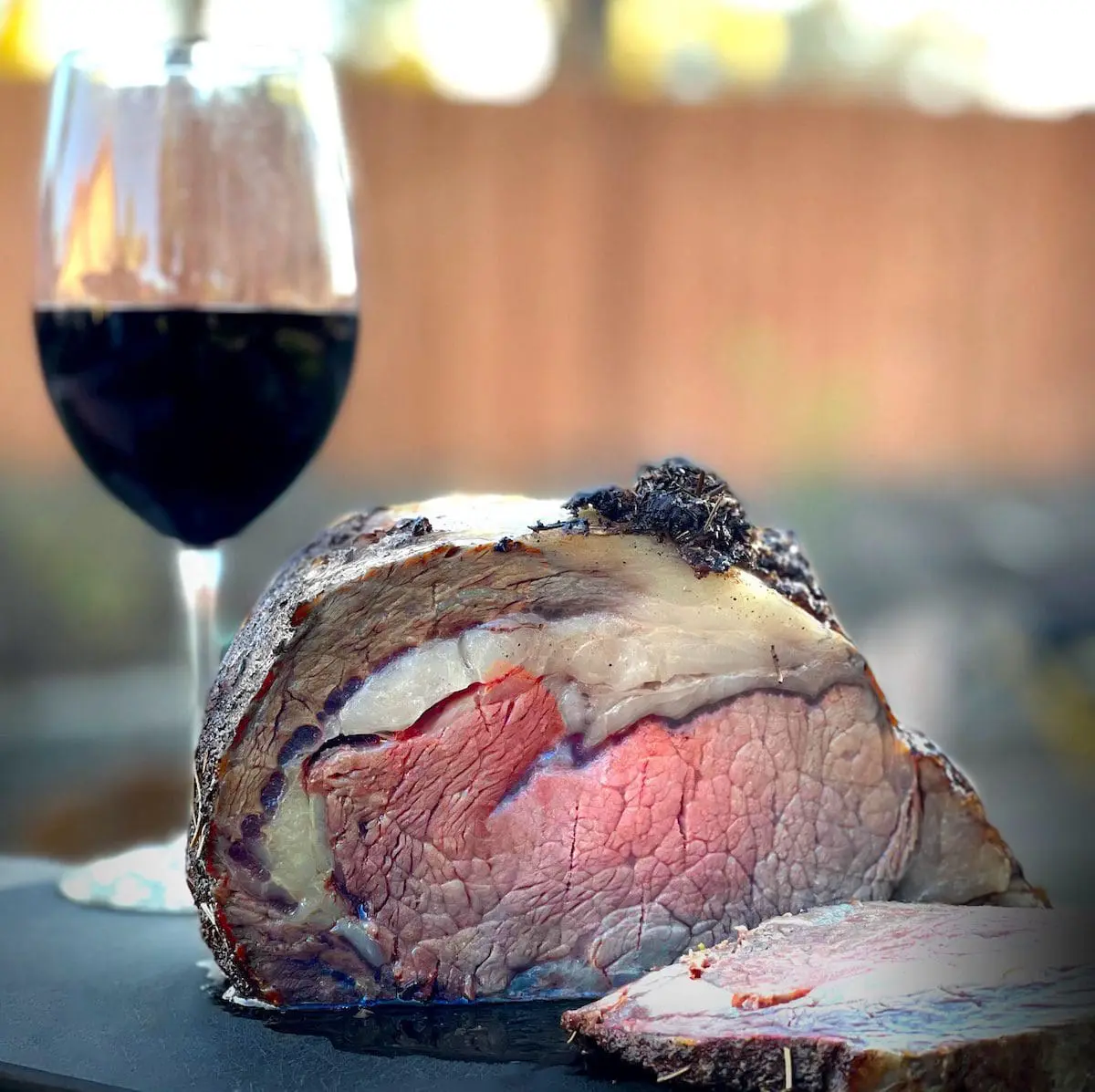 A smoked prime rib roast with one slice cut off laying on a black cutting board. A glass of red wine is setting next to the cutting board.