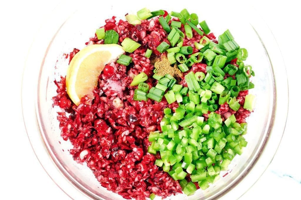 Ingredients to make cranberry jalapeno dip in a clear mixing bowl: cranberries, jalapenos, green onions and lemon.
