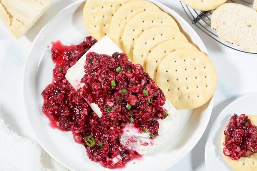 a block of cream cheese topped with Cranberry jalapeno dip. Butter crackers and bread slices are in the background.