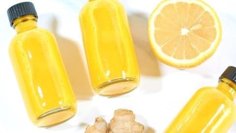 Photo of three clear glass bottles filled with lemon and ginger shots. A knob of ginger and half a slice of lemon are next to the bottles.
