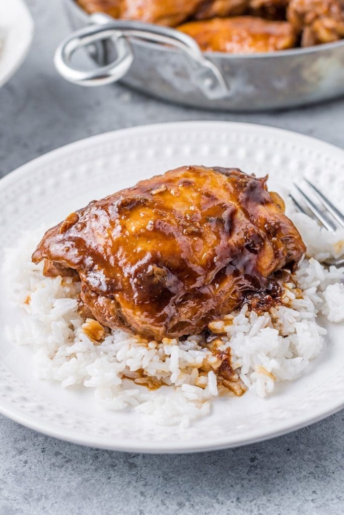 A white plate with cooked Chicken placed upon a bed of white rice.