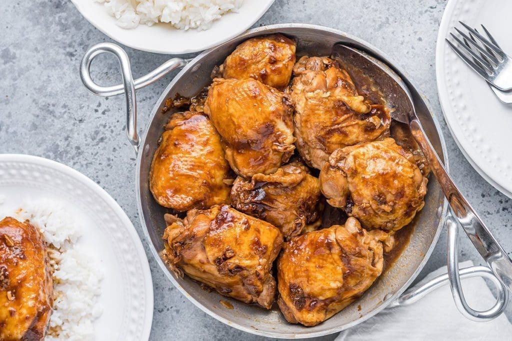 Filipino Chicken plated on a silver platter surrounded by white rice and white plates and forks.