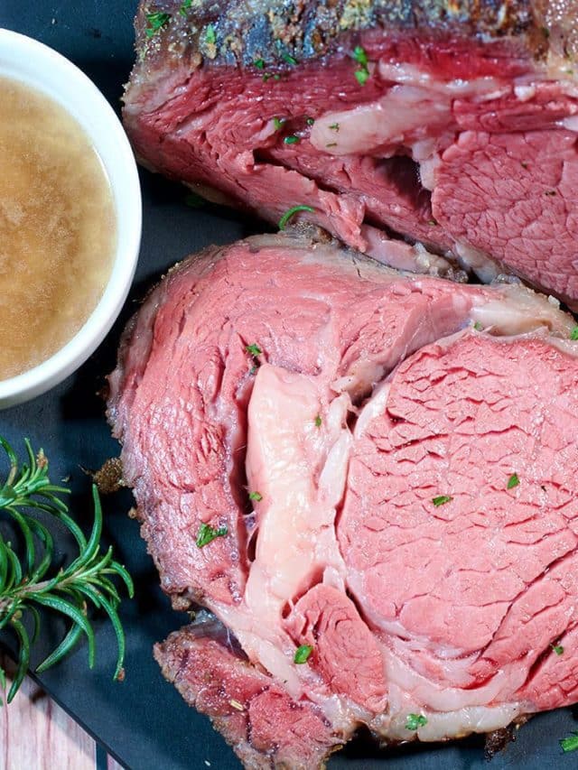 Close up picture of rare prime rib sliced on a black cutting board. Au jus is next to it in a white cup.