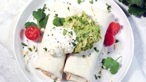 Two frozen burritos in air fryer topped with guacamole, sour cream, tomatoes and cilantro on a white plate.