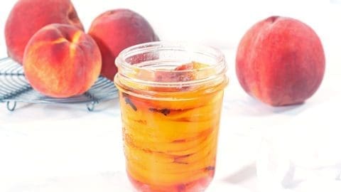 Pint size mason jar filled with pickled peaches resting on a white napkin. Four whole fresh peaches are in the background.