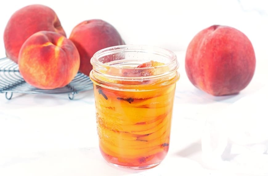 Pint size mason jar filled with pickled peaches resting on a white napkin. Four whole fresh peaches are in the background.