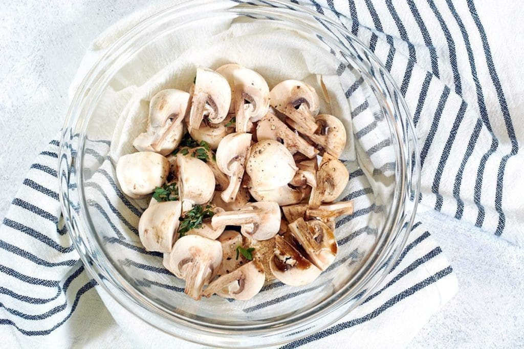 Close-up of marinated mushrooms in a clear mixing bowl sitting on a peeled napkin.