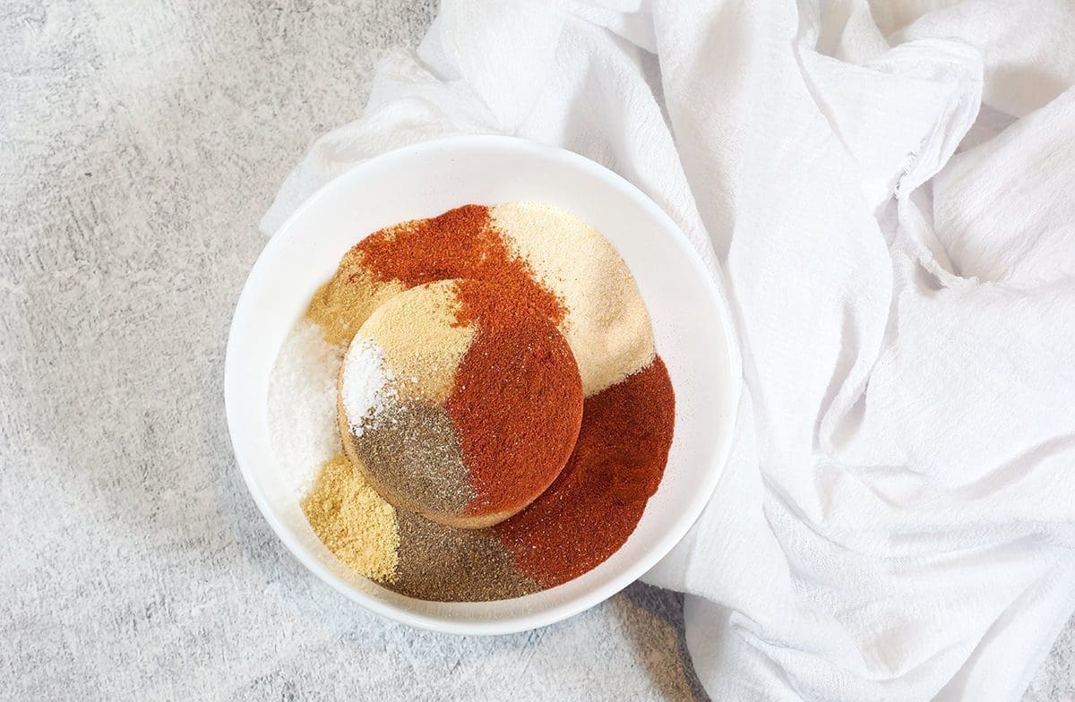 Individual spices needed to make BBQ Rub all spaced out in a white bowl next to a white cloth napkin.