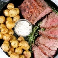 Top down view of a smoked beef tenderloin roast cut into 5 medallions. Served alongside baby potatoes and horseradish sauce on a black cat iron pan.
