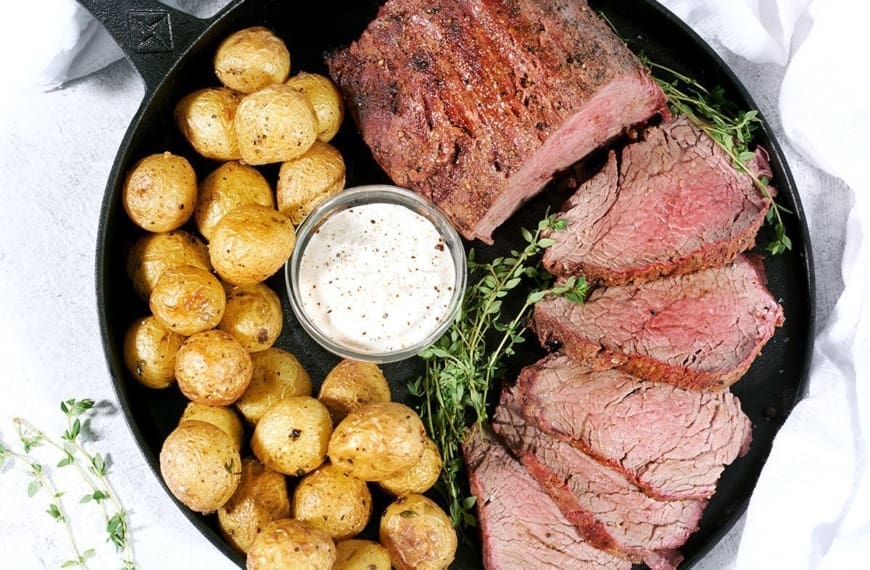 Top down view of a smoked beef tenderloin roast cut into 5 medallions. Served alongside baby potatoes and horseradish sauce on a black cat iron pan.