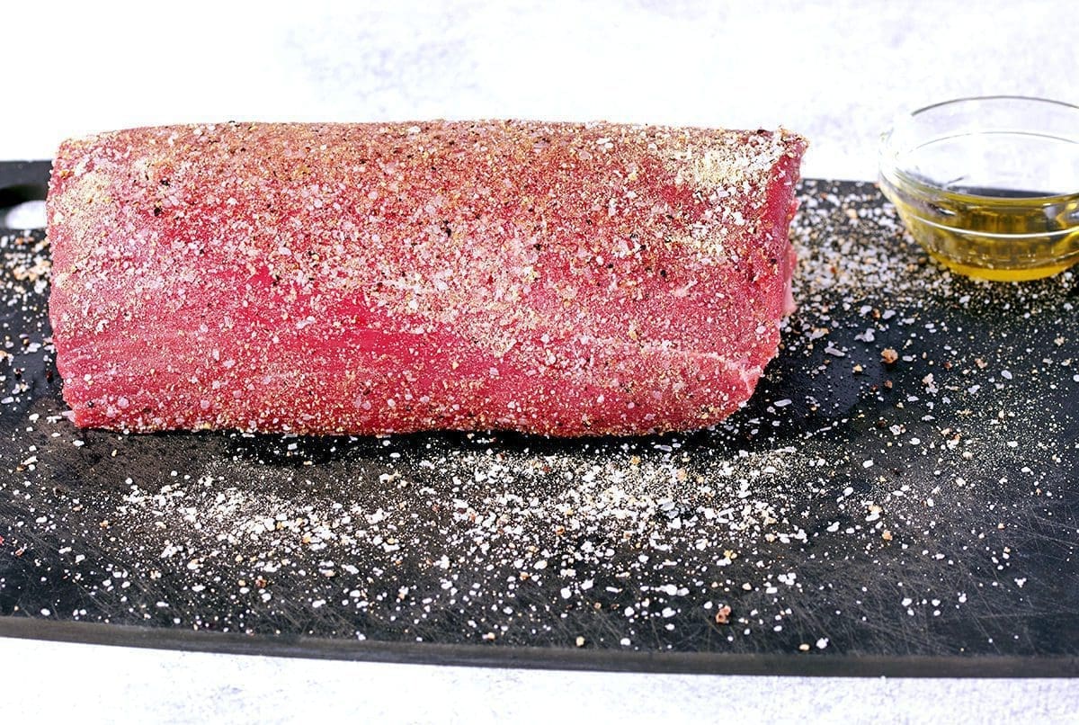 Side view of an uncooked roast covered in seasonings. A clear cup of oil is nearby.