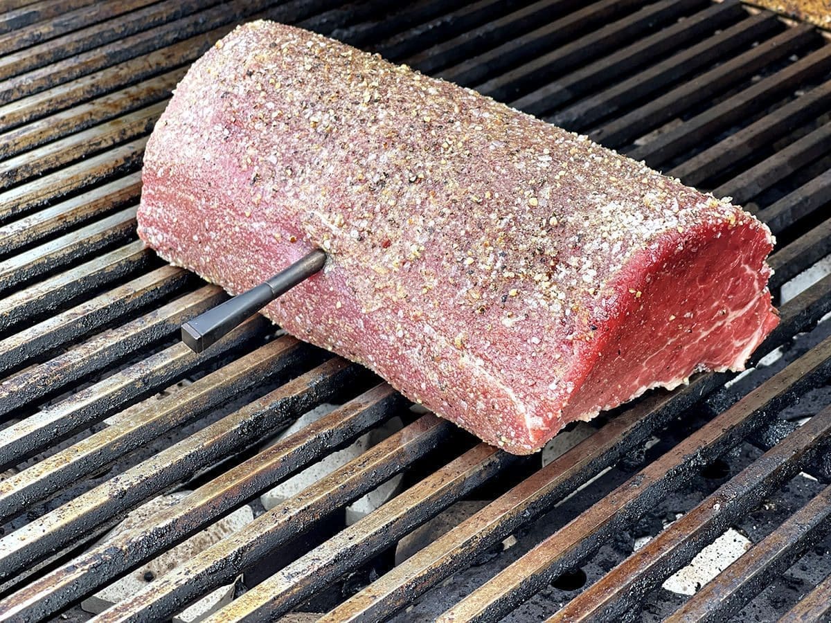 Side view of a rare beef roast on a grill. A metal thermometer is sticking out of the side.