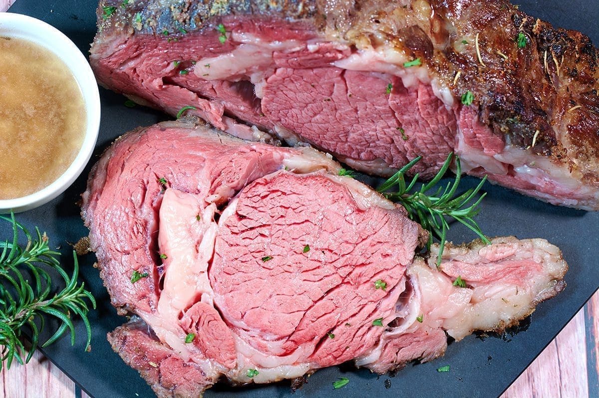 Up close shot of sliced sous vide prime rib, on a black cutting board, with a side of au jus in a white glass cup.