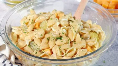 Clear bowl of mixed dill pickle pasta salad with extra cheese, pickles and onions on the side.