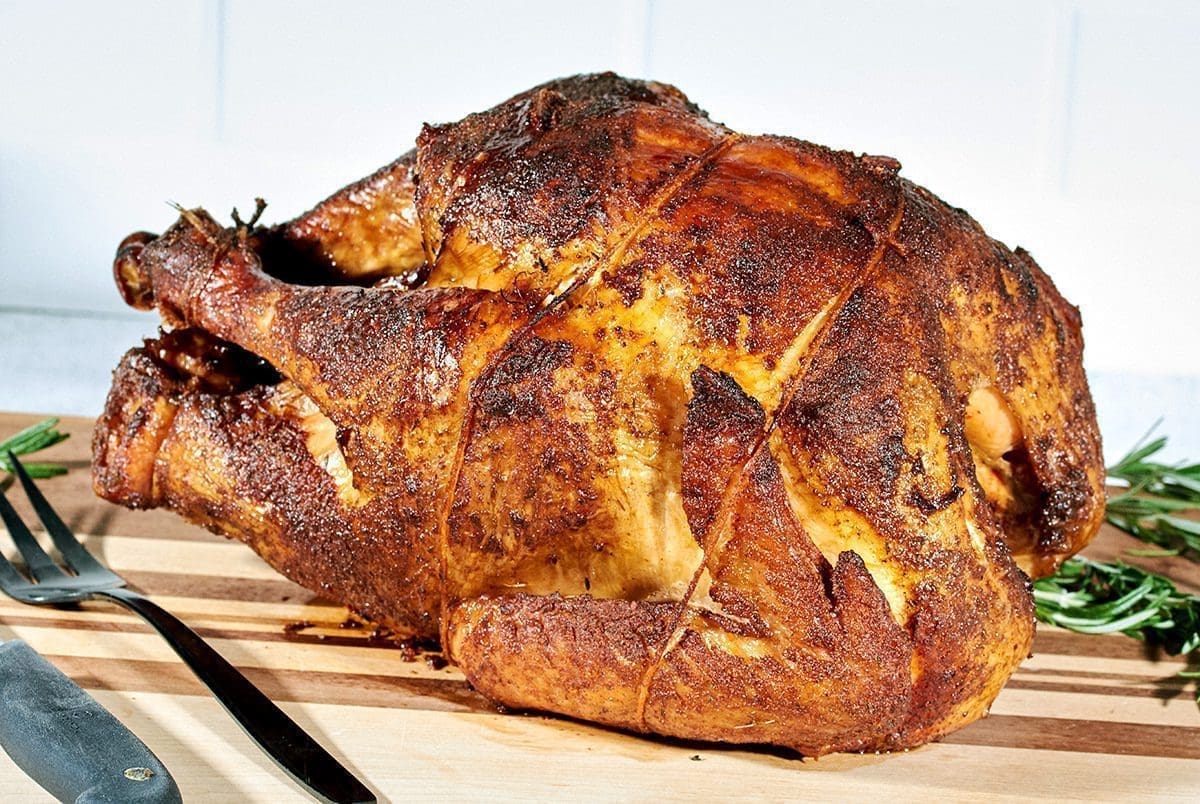 Close up of a cooked smoked turkey on a striped cutting board. A knife and large fork are next to the bird.