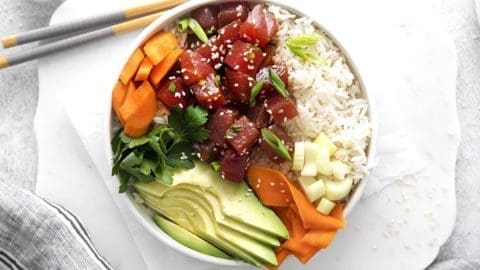 Overheard shot of an ahi tuna poke bowl, including tuna, avocado, carrots and rice. Chop sticks and a white napkin are next to the bowl.