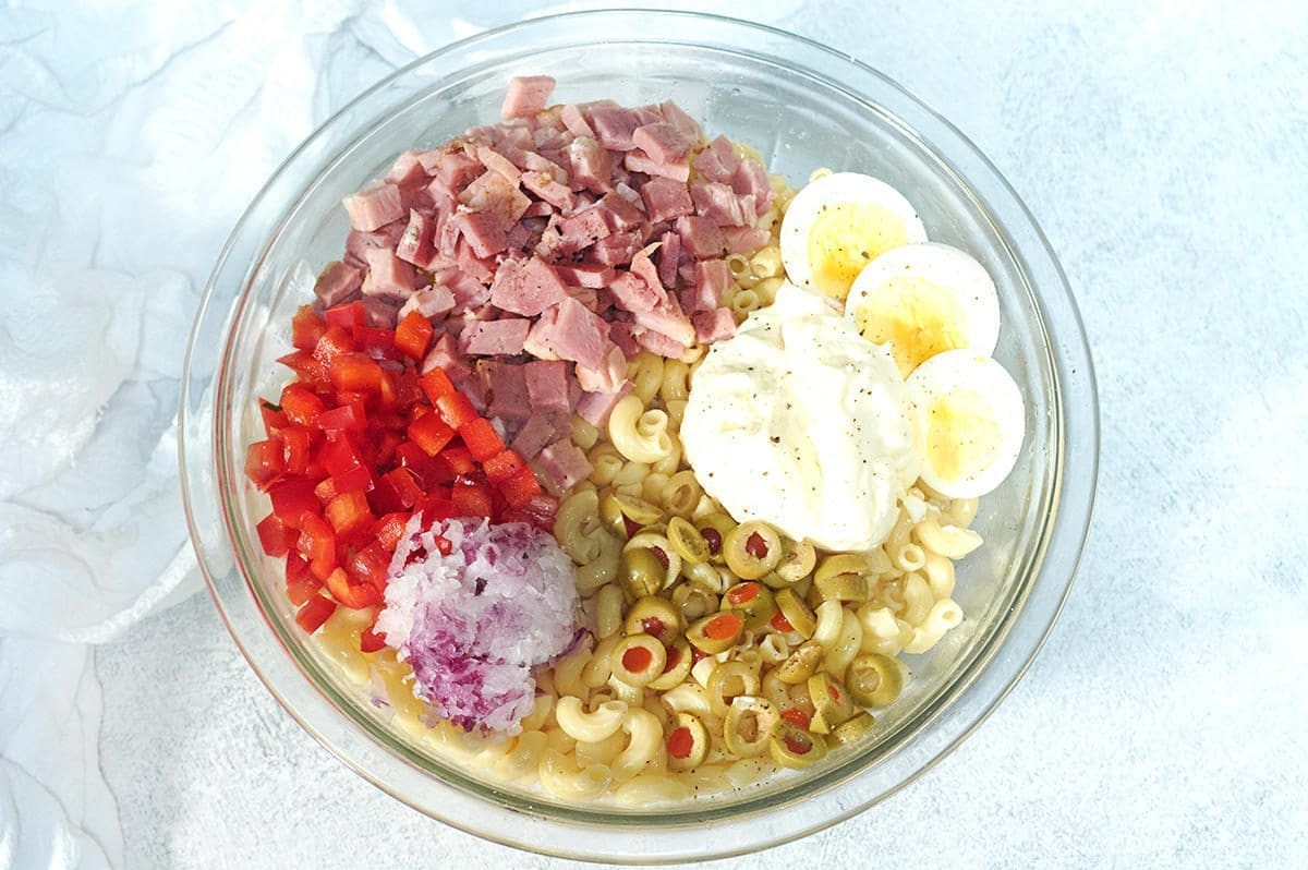 Ingredients for ensalada de coditos in a large clear bowl surrounded by a white napkin.