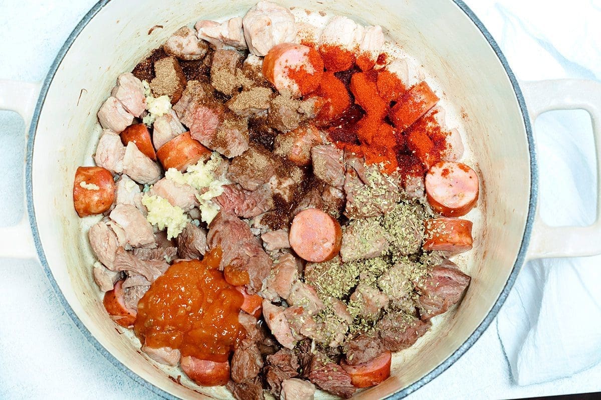 Browned cubed pork, beef and sausage mixed with onions and bell peppers in a cream dutch oven. Sofrito and herbs are sprinkled on top of the browned meat.