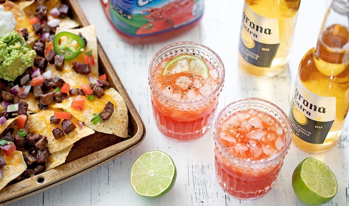 Two glasses of Michelada next to sheet pan nachos with 4 ingredient guacamole, lime wedges and Corona bottles.