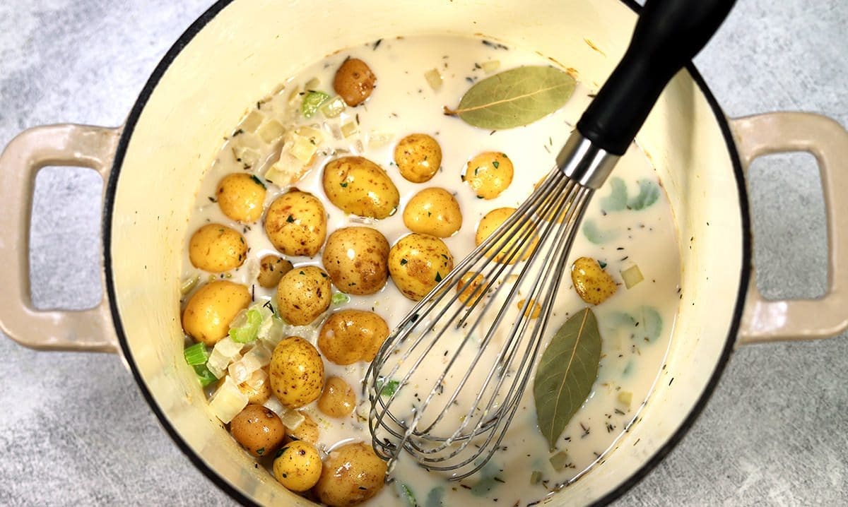 Dutch oven with tiny potatoes, bay leaves and cream. A black whisk is set inside the dutch oven.