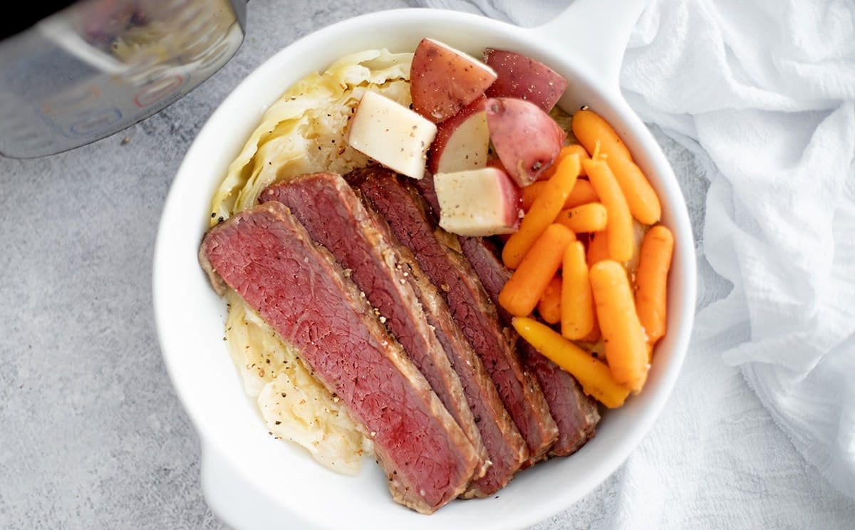 Instant Pot Corned Beef, carrots, cabbage and potatoes in a white bowl. An instant pot and white napkin are next to it.
