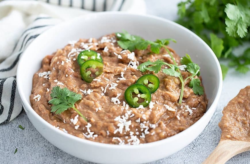 Instant Pot Refried Beans in a white bowl topped with cheese, jalapenos and cilanto.