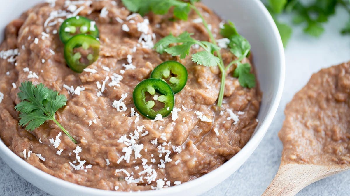 Close up of refried beans in a white bowl topped with jalapenos, cheese and cilantro.
