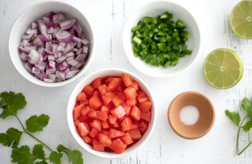 In a white bowl, diced tomatoes, onions and jalapenos are diced in a white bowl with lime wedges, salt and cilantro next to it.