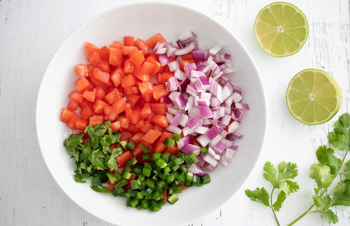Diced tomatoes, onions and jalapenos in one white bowl with lime wedges, salt and cilantro next to them.