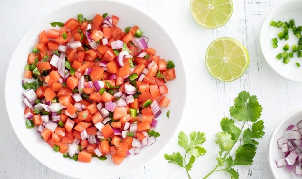 Pico de gallo in a white bowl surrounded by individual ingredients.