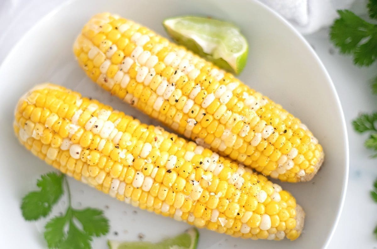 Two ears of corn on a white plate accompanied by cilantro and lime wedge.