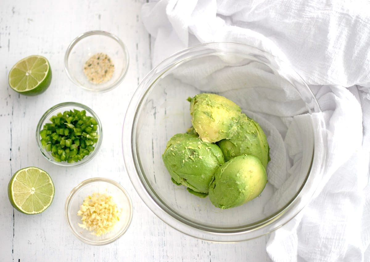 Clear mixing bowls with avocado, jalapeno, salt and garlic in them. A cut lime and white napkin are next to the bowls.
