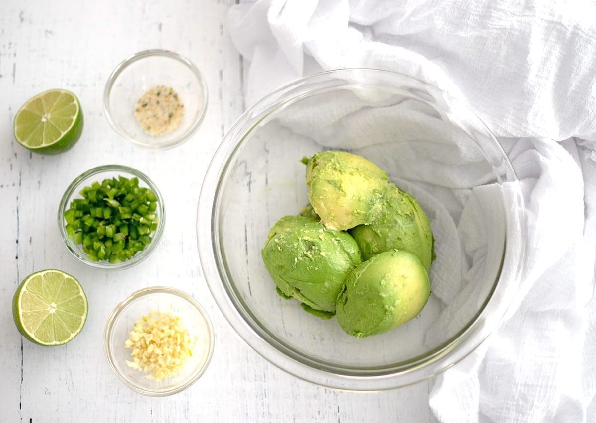 Clear the mixing bowl with avocado, jalapeno, salt and garlic. Cut lime and white napkins are located next to the bowl.
