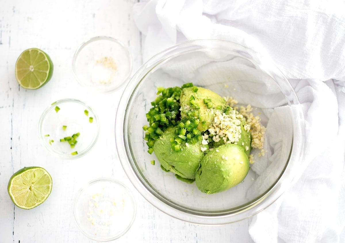 a large mixing bowl with avocado, jalapeno and garlic unmixed. Empty small mixing bowls and a lime are nearby.