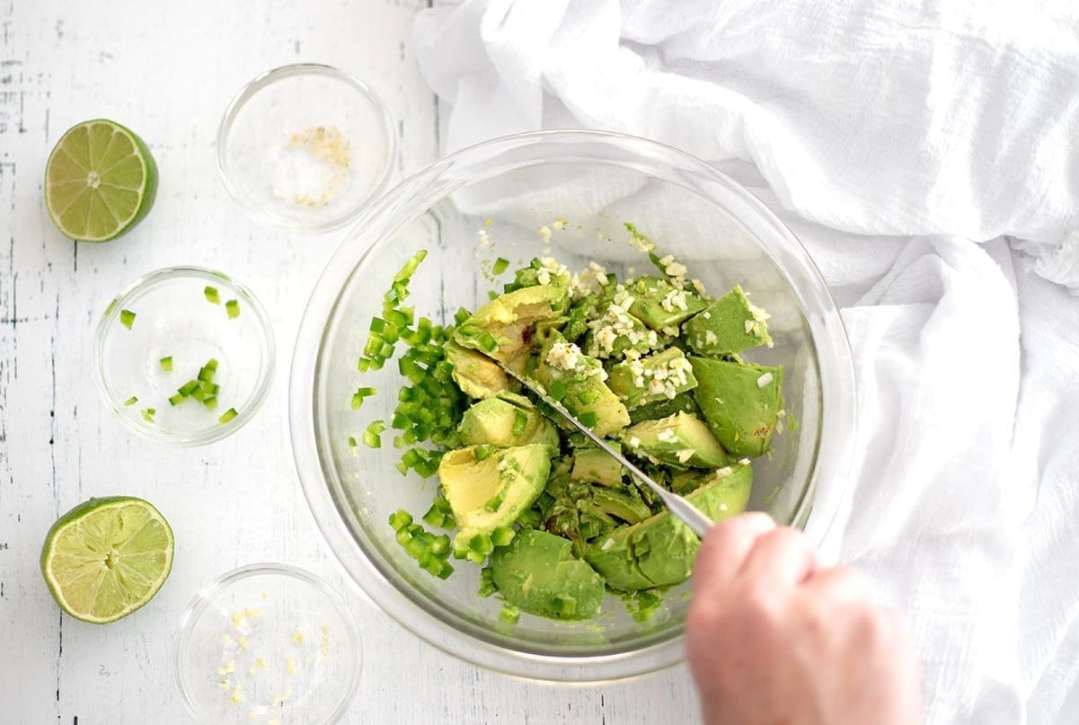 A large mixing bowl of avocado, jalapeno and garlic mixed with butter knife. A small empty mixing bowl and lime are nearby.