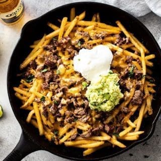 Carne Asada Fries in a black cast iron skillet with a corona, bowl of salsa and slices of lime next to it.