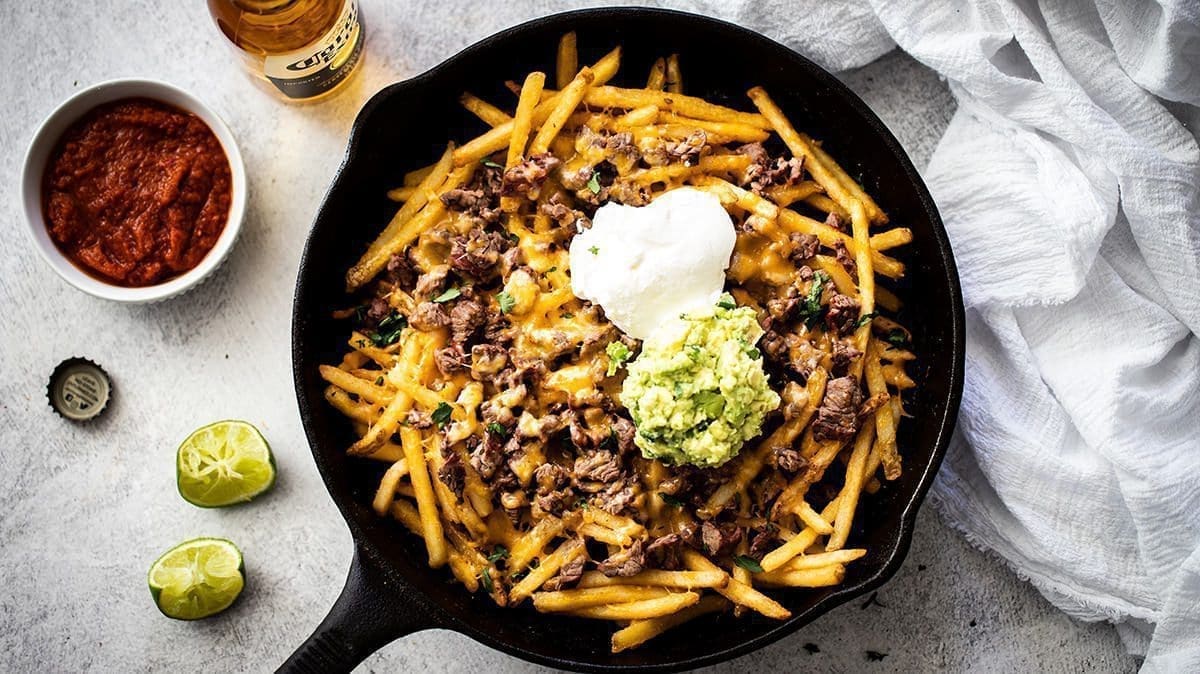 Carne Asada Fries in a black cast iron skillet with a corona, bowl of salsa and slices of lime next to it.