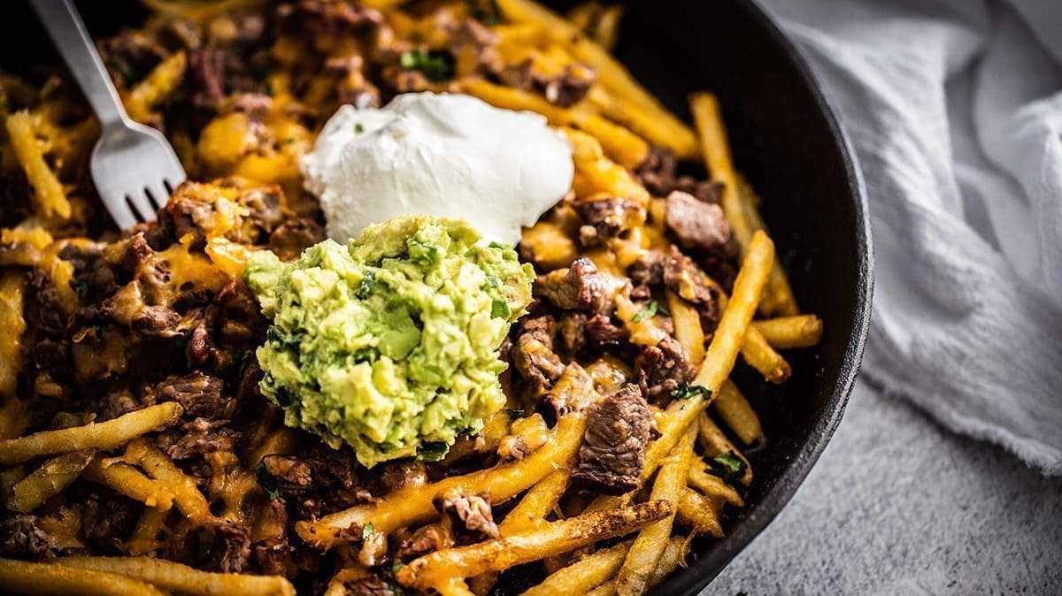 Close up of french fries with steak, guacamole and sour cream on them in a black skillet.