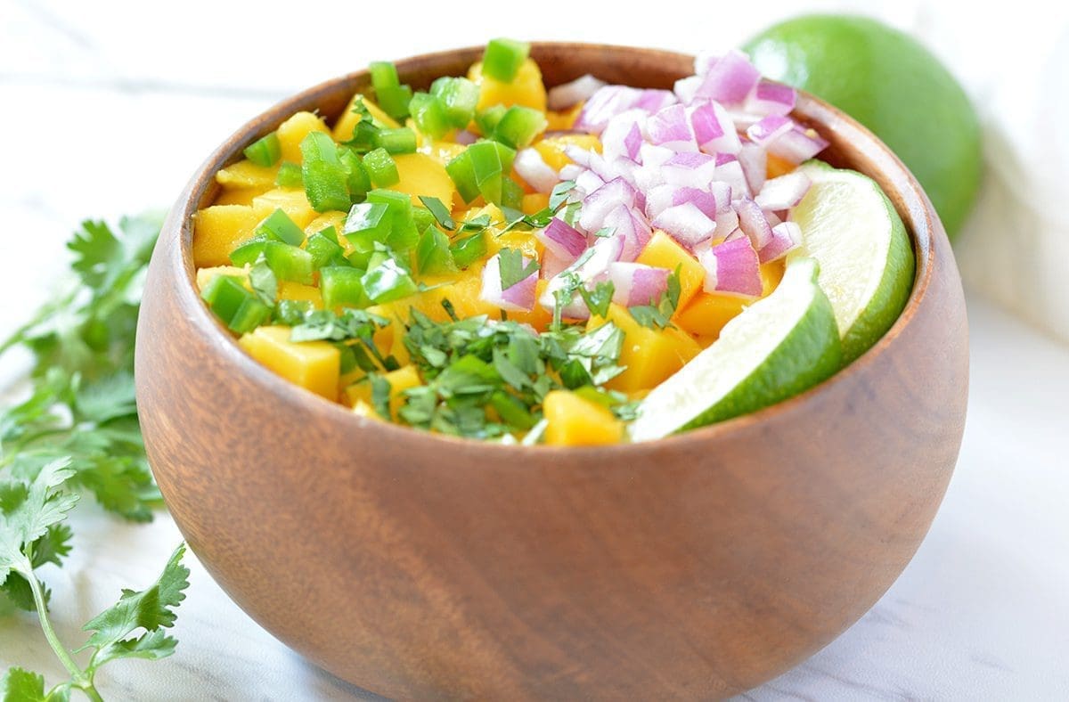 Side view of a wooden bowl with diced mango, jalapenos, onions and lime slices inside. A sprig of cilantro is off to the side.