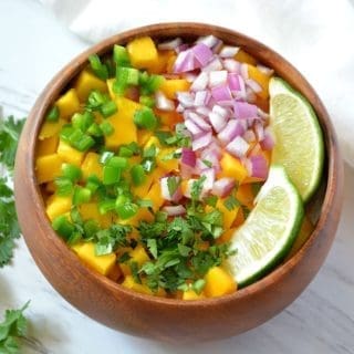 Wooden bowl with diced mango, jalapenos, onions and lime slices inside. A sprig of cilantro is off to the side.